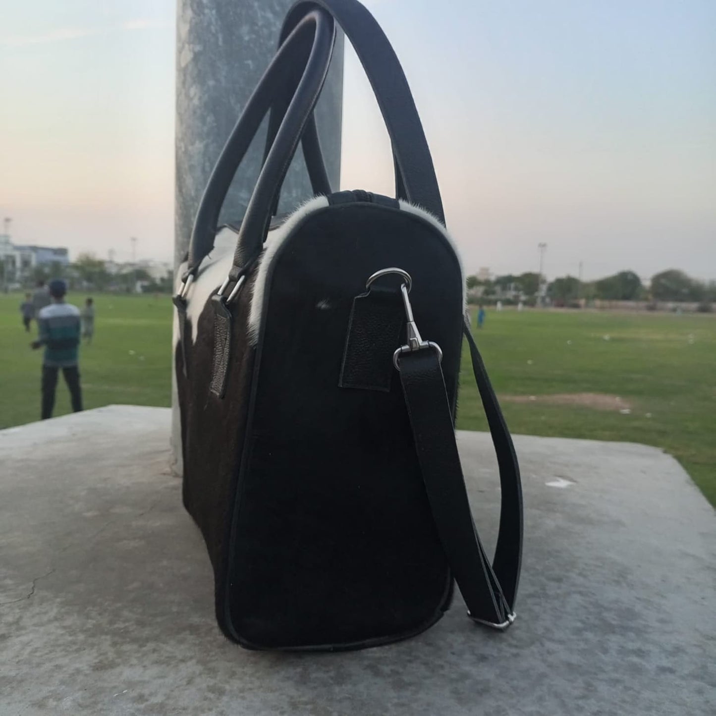 Cowgirl wearing denim with cowhide bag, standing in a field at sunset.