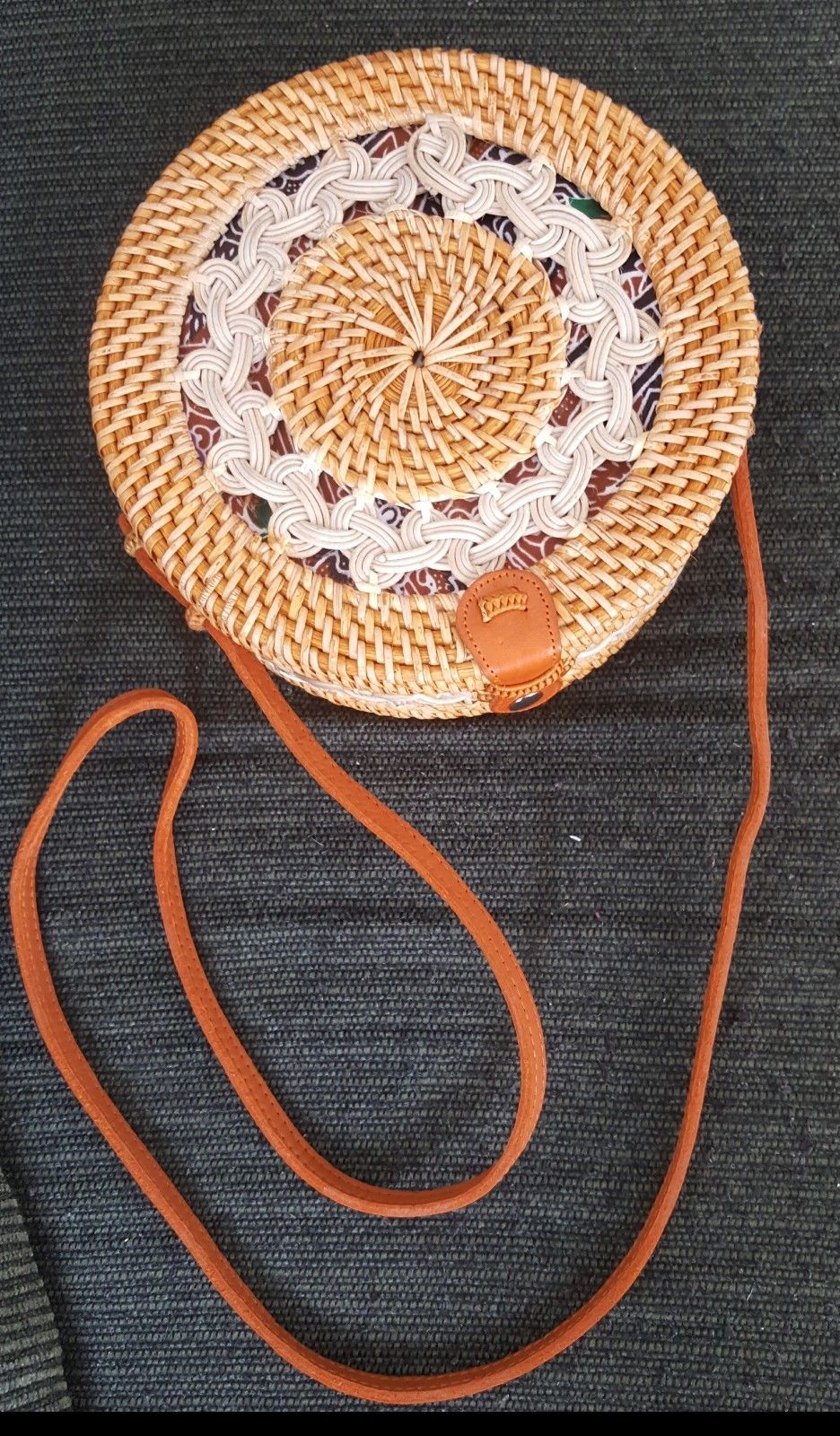 Handwoven rattan purse with a circular design, adorned with a leather strap, resting on a light, textured surface.