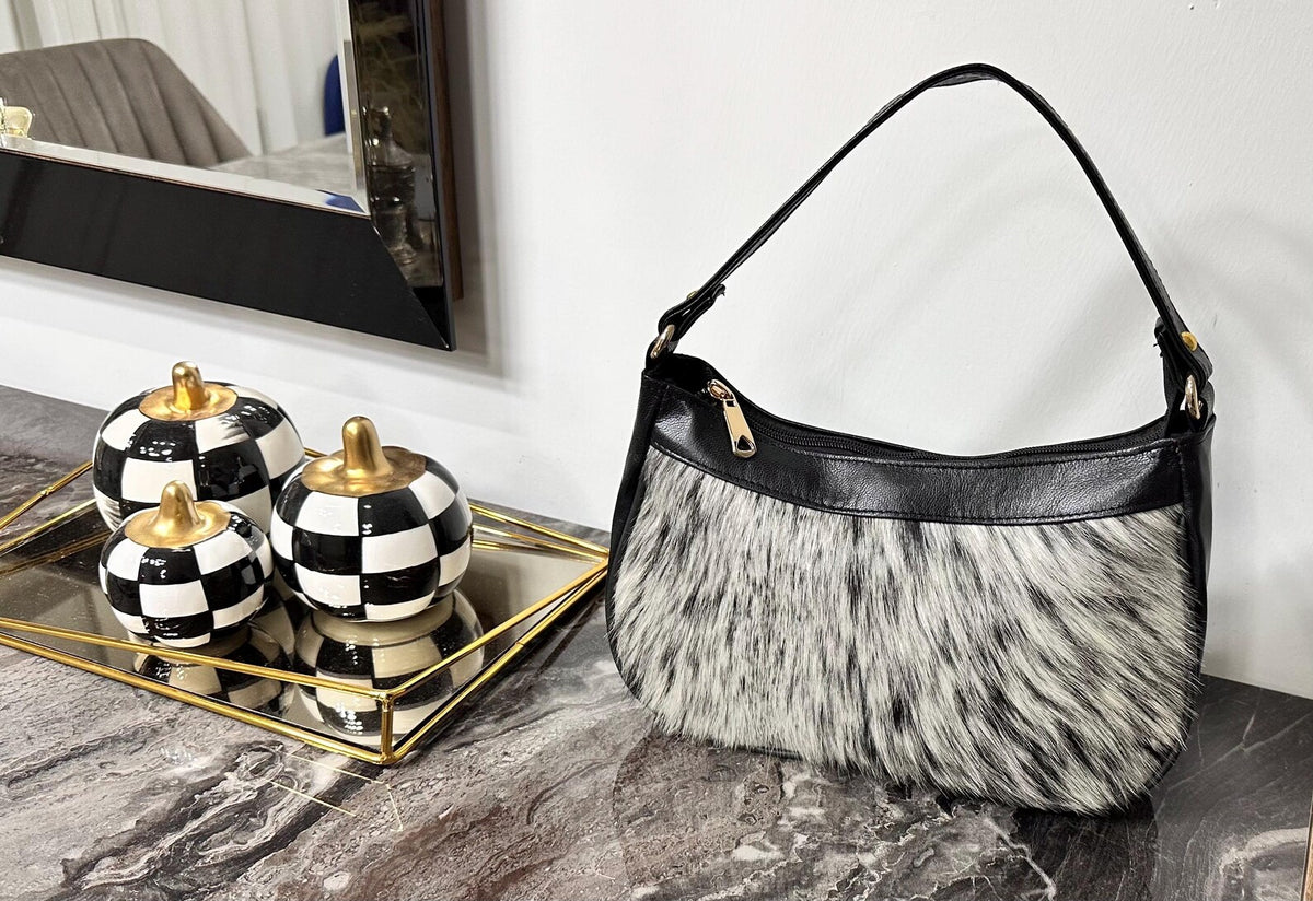Cowhide bag with natural gray and black hide variation, zip-top opening, leather trim, and compact shoulder-carry shape.
