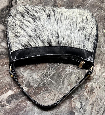 Cowhide bag with slim structured profile, black shoulder strap, gold-tone hardware, and natural hair-on cowhide patterning.