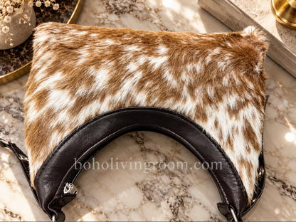 Cowhide shoulder bag with curved hobo-inspired silhouette, black shoulder strap, white markings, and warm camel-toned hide.