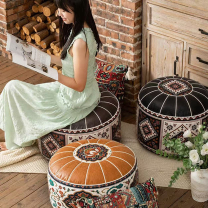 Three decorative round poufs in a rustic room setting, with a woman in a light green dress sitting on one of the poufs and reading a magazine. The poufs feature intricate geometric and tribal patterns in black, brown, red, and cream, with one pouf in the foreground showing a warm brown leather top.