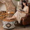 A woman in a beige dress sits in a brown leather armchair reading a magazine, with her foot resting on a round decorative ottoman. The room has a rustic brick fireplace, wood logs, and a small wooden side table with a cup.