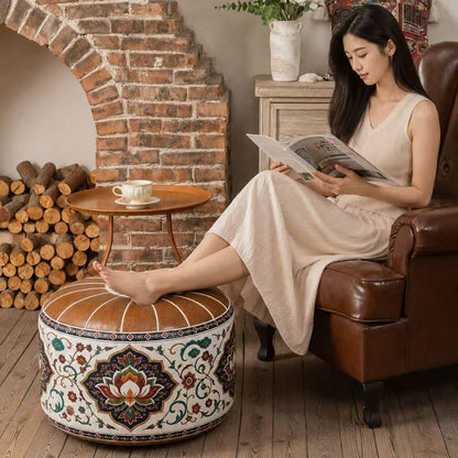 A woman in a beige dress sits in a brown leather armchair reading a magazine, with her foot resting on a round decorative ottoman. The room has a rustic brick fireplace, wood logs, and a small wooden side table with a cup.