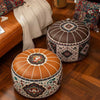 Two round Moroccan-style pouf ottomans on a wooden floor in a styled living room. The foreground ottoman is made of tan leather with white radial stitching and a multicolor floral medallion on top, while the background ottoman is dark brown with white stitching and a geometric floral pattern on the sides.
