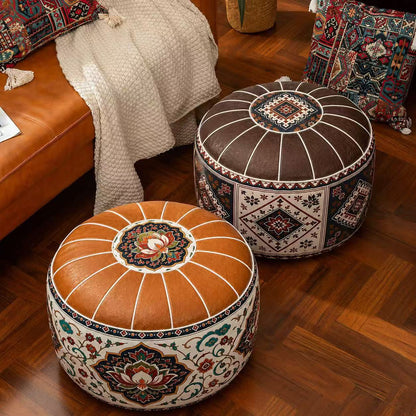 Two round Moroccan-style pouf ottomans on a wooden floor in a styled living room. The foreground ottoman is made of tan leather with white radial stitching and a multicolor floral medallion on top, while the background ottoman is dark brown with white stitching and a geometric floral pattern on the sides.