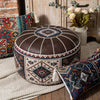 A round brown faux-leather pouf ottoman with a bohemian-style geometric pattern, styled in a rustic room setting with other patterned pillows and a vase of flowers.