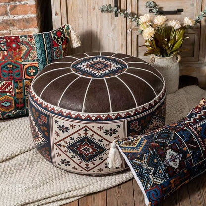 A round brown faux-leather pouf ottoman with a bohemian-style geometric pattern, styled in a rustic room setting with other patterned pillows and a vase of flowers.