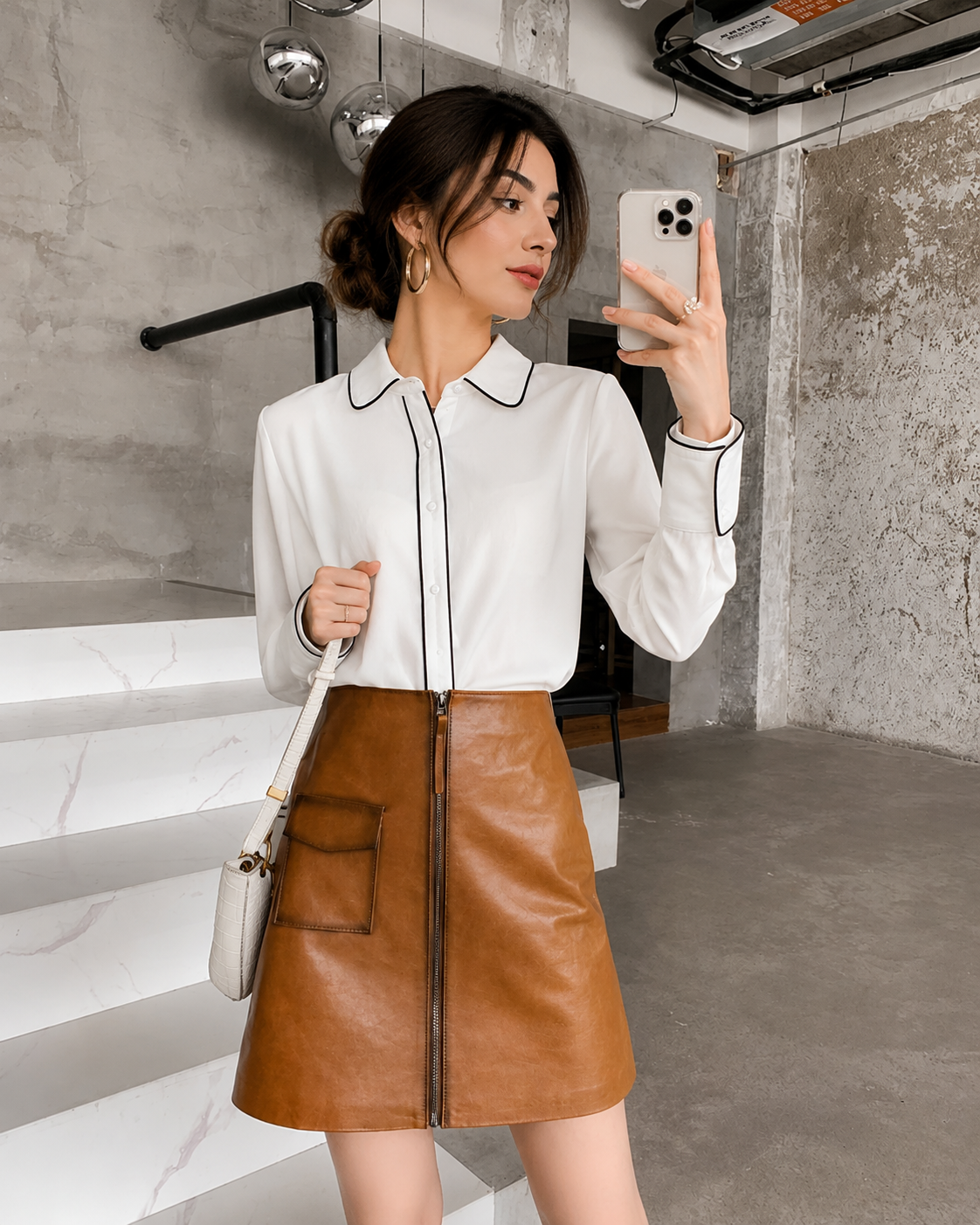 Woman taking a mirror selfie wearing a white blouse and brown leather skirt.
