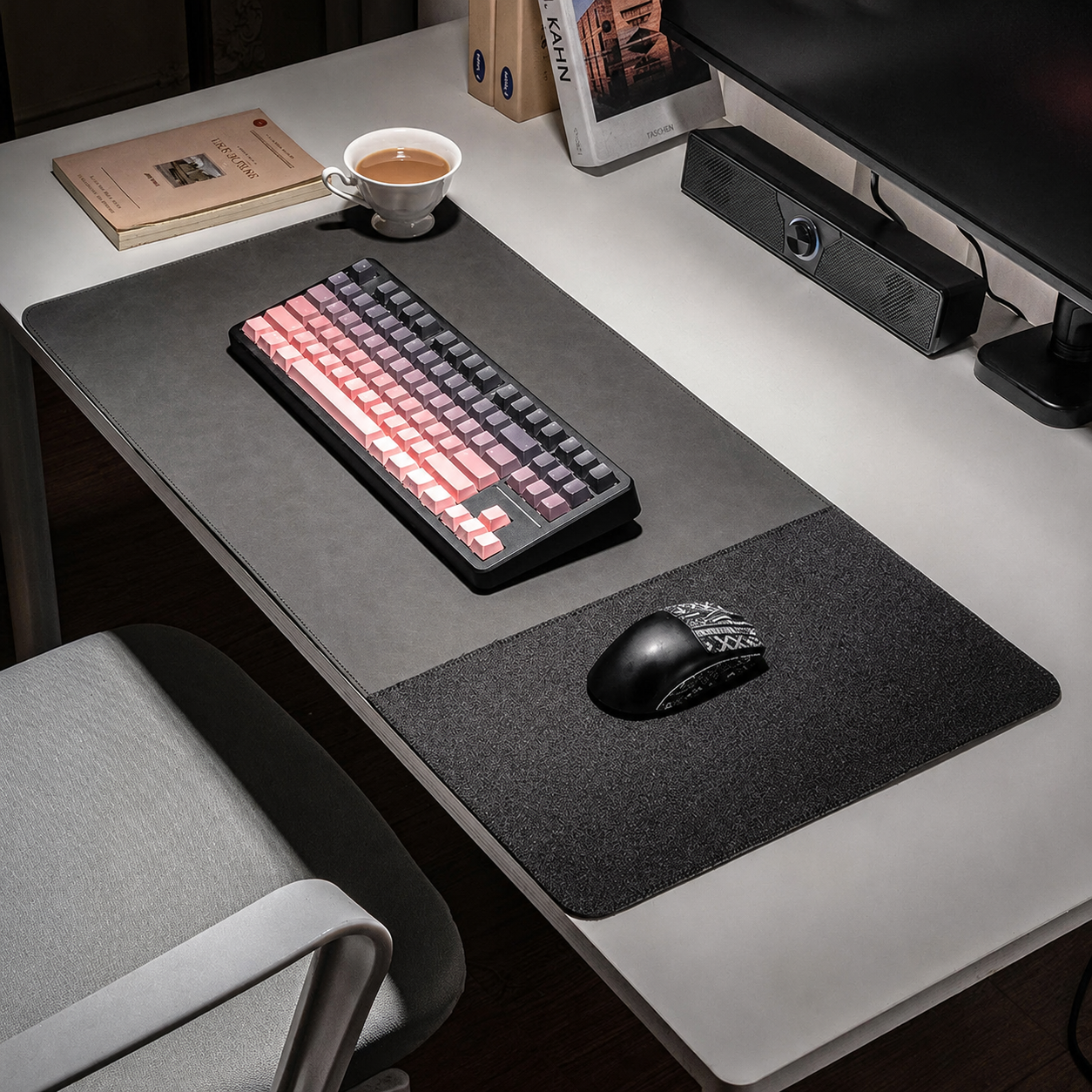 A large dark gray desk mat with a built-in wrist rest is laid out on a white desk, styled with a mechanical keyboard, a black computer mouse, a cup of coffee, and other desk accessories.