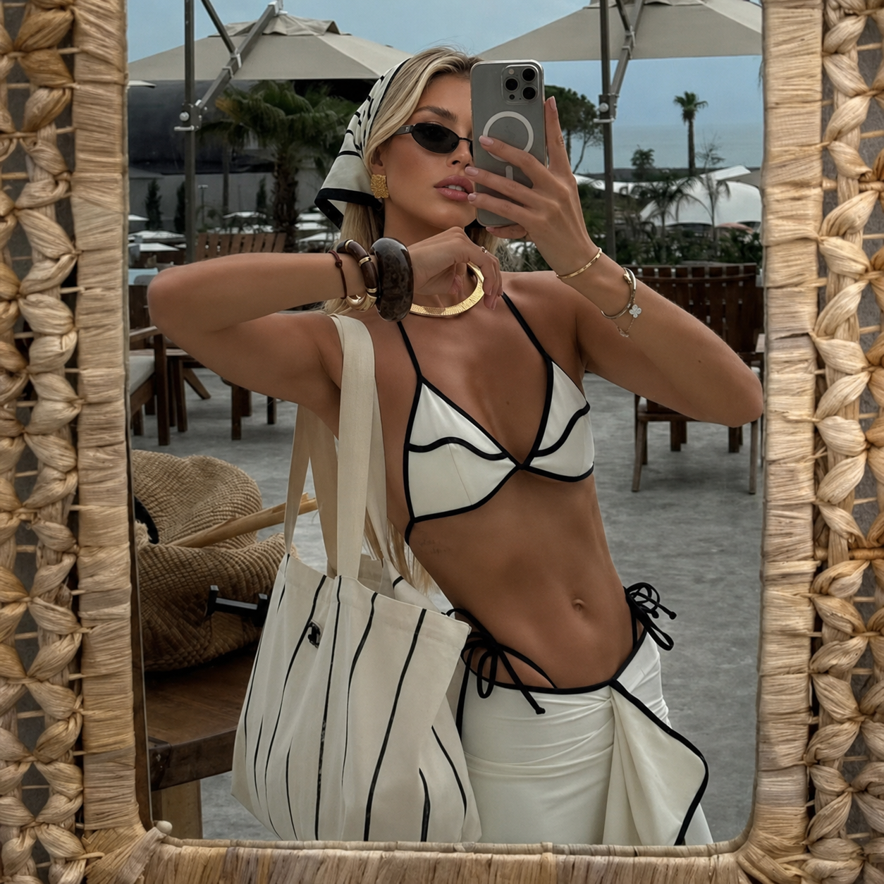 A woman takes a mirror selfie in an outdoor patio setting, wearing a white bikini with black trim and matching white bottoms, carrying a large cream tote bag with black stripes.