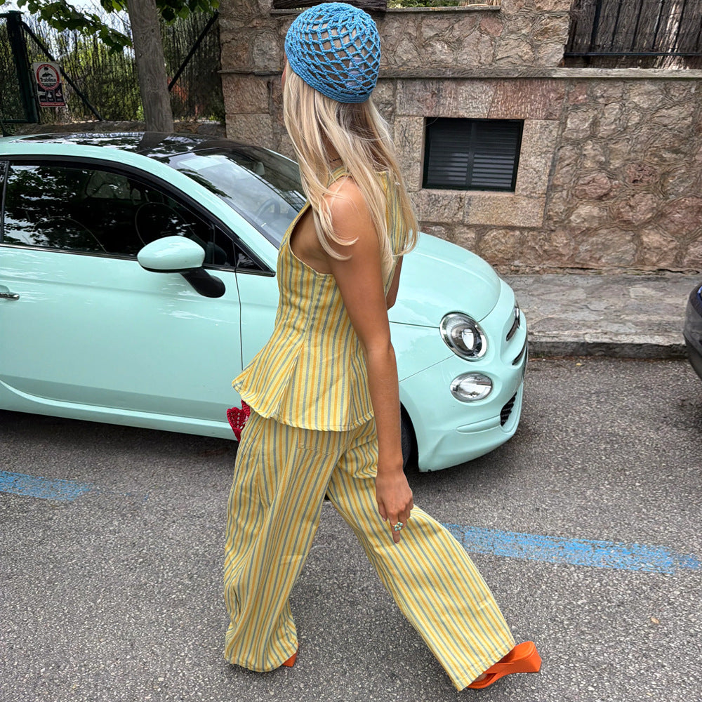 A woman wearing a yellow and white striped sleeveless top and matching wide-leg pants, a blue crochet beanie, and orange shoes, walking in front of a mint green car and a stone wall.