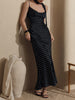 A woman models a black halter-neck maxi dress with a white polka dot pattern, styled with a matching choker and black sandals in a warm, minimalist interior setting.
