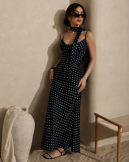 A woman wearing a black halter-neck maxi dress with white polka dots, standing in a minimalist room with textured beige walls and a wooden chair.