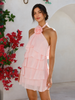A woman models a light pink halter mini dress with a tiered ruffle skirt and a large fabric flower detail at the neckline, set against a white stucco wall with red flowers on the left.