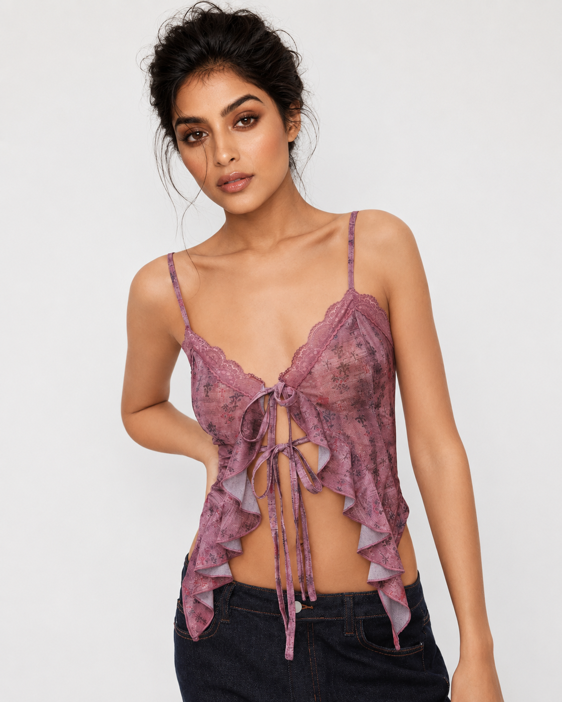 A woman modeling a dusty purple lace camisole top with thin straps, ruffled edges, and a front tie closure, paired with dark jeans against a plain light gray studio background.