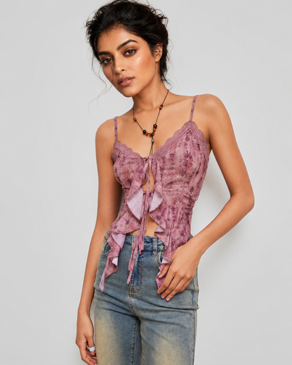 A woman modeling a mauve pink lace corset-style camisole top with ruffle details and tie-front keyhole, paired with blue jeans against a light gray studio background.