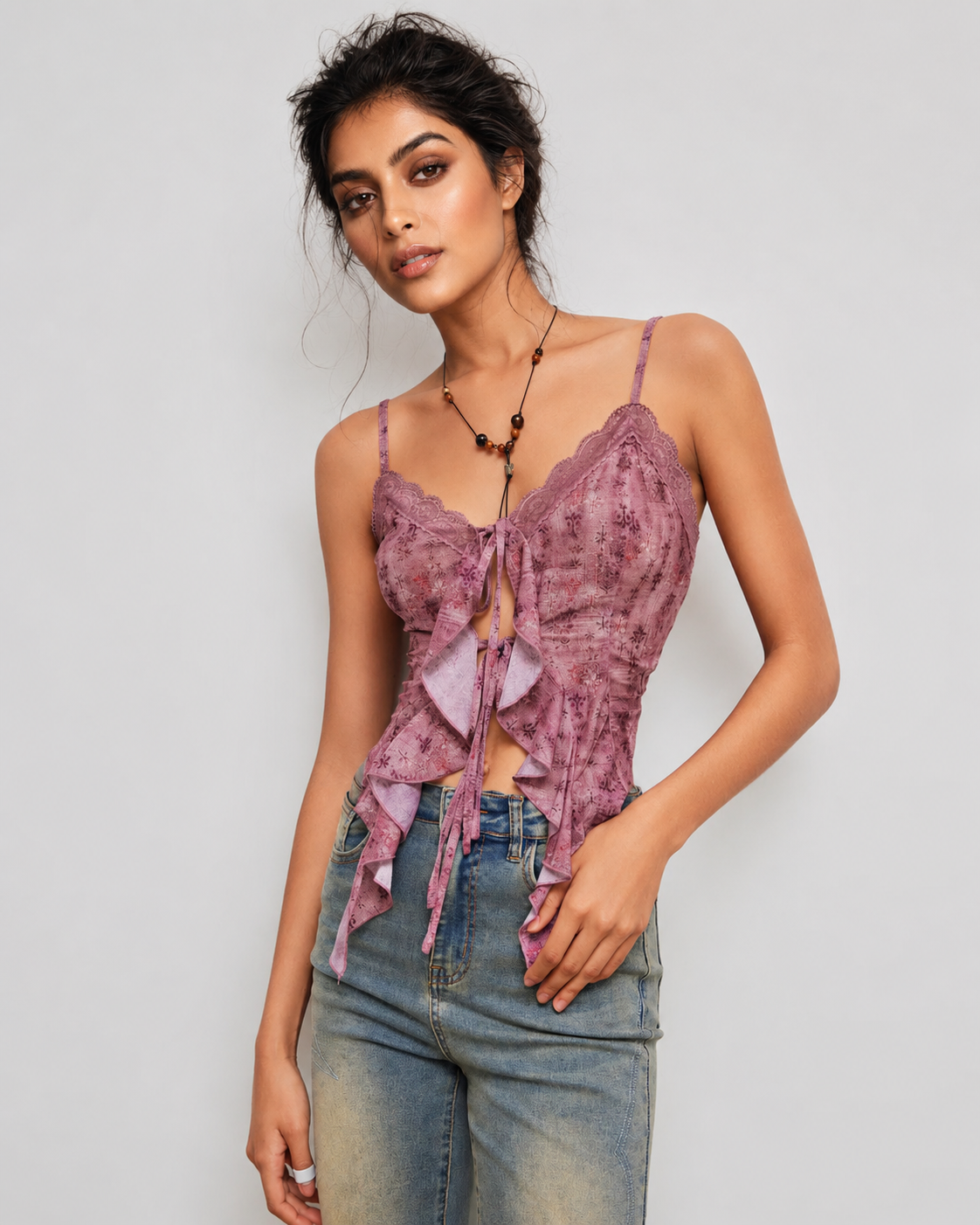 A woman modeling a mauve-purple lace camisole top with ruffle details and tie-front closures, paired with light-wash blue jeans against a light gray studio background.