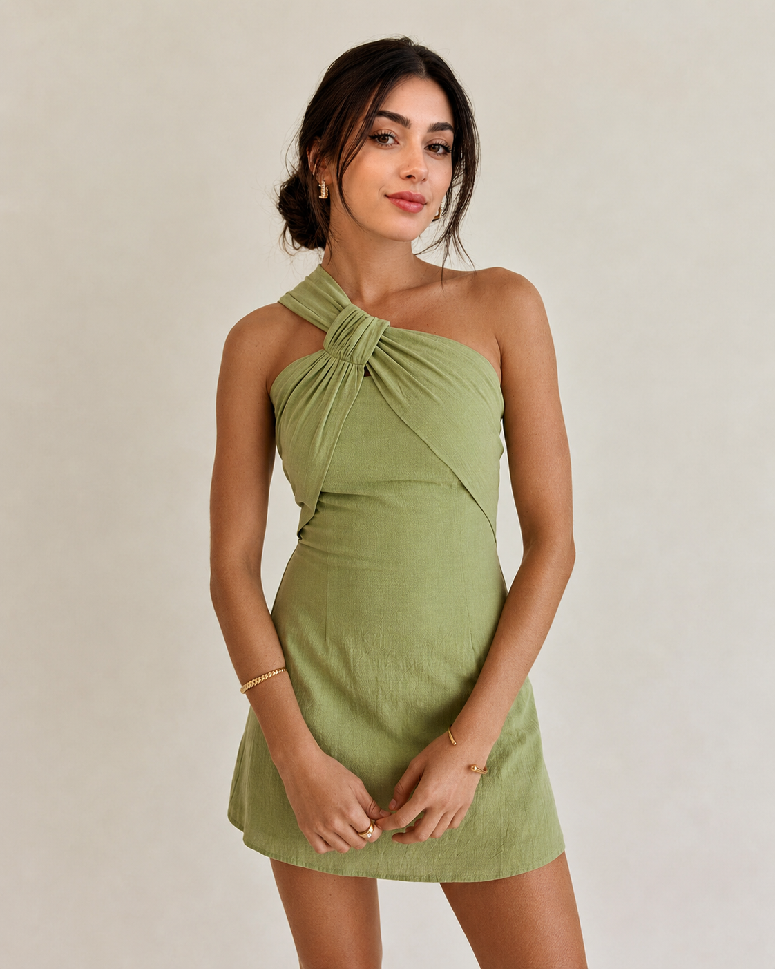 A woman modeling a light olive green one-shoulder mini dress with a knotted crossover detail at the neckline, set against a plain light beige studio background.