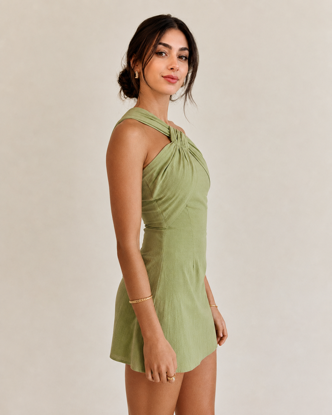 A woman modeling a sage green halter mini dress with a twisted knot detail at the neckline, set against a plain beige studio background.