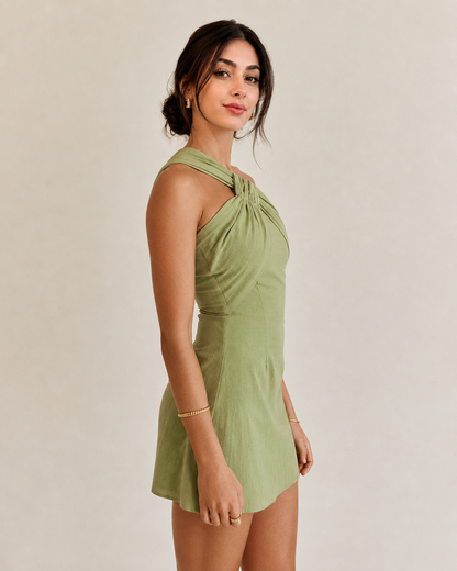 A woman modeling a sage green halter mini dress with a twisted knot detail at the neckline, set against a plain beige studio background.