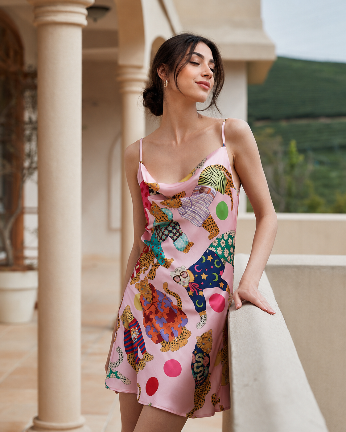 A woman wearing a pink satin slip dress with a colorful patchwork print of animals and dots, posing on a sunlit outdoor terrace with beige columns.