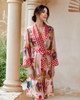 A woman wearing a pink kimono-style robe with red and white polka dot trim and a multicolor animal print, standing in front of a beige stone wall with columns.