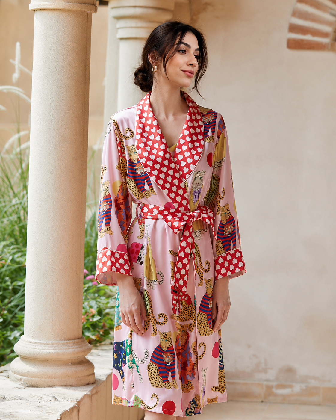 A woman wearing a pink kimono-style robe with red and white polka dot trim and a multicolor animal print, standing in front of a beige stone wall with columns.