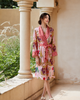 A woman wearing a pink and red patterned kimono robe with a red and white polka dot collar and waist tie, standing on a beige stone porch with columns.