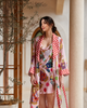 A woman wearing a pink patterned robe and matching dress stands in a warm-toned architectural setting with columns and a tree, featuring a multicolor print with red polka dots and whimsical illustrations.
