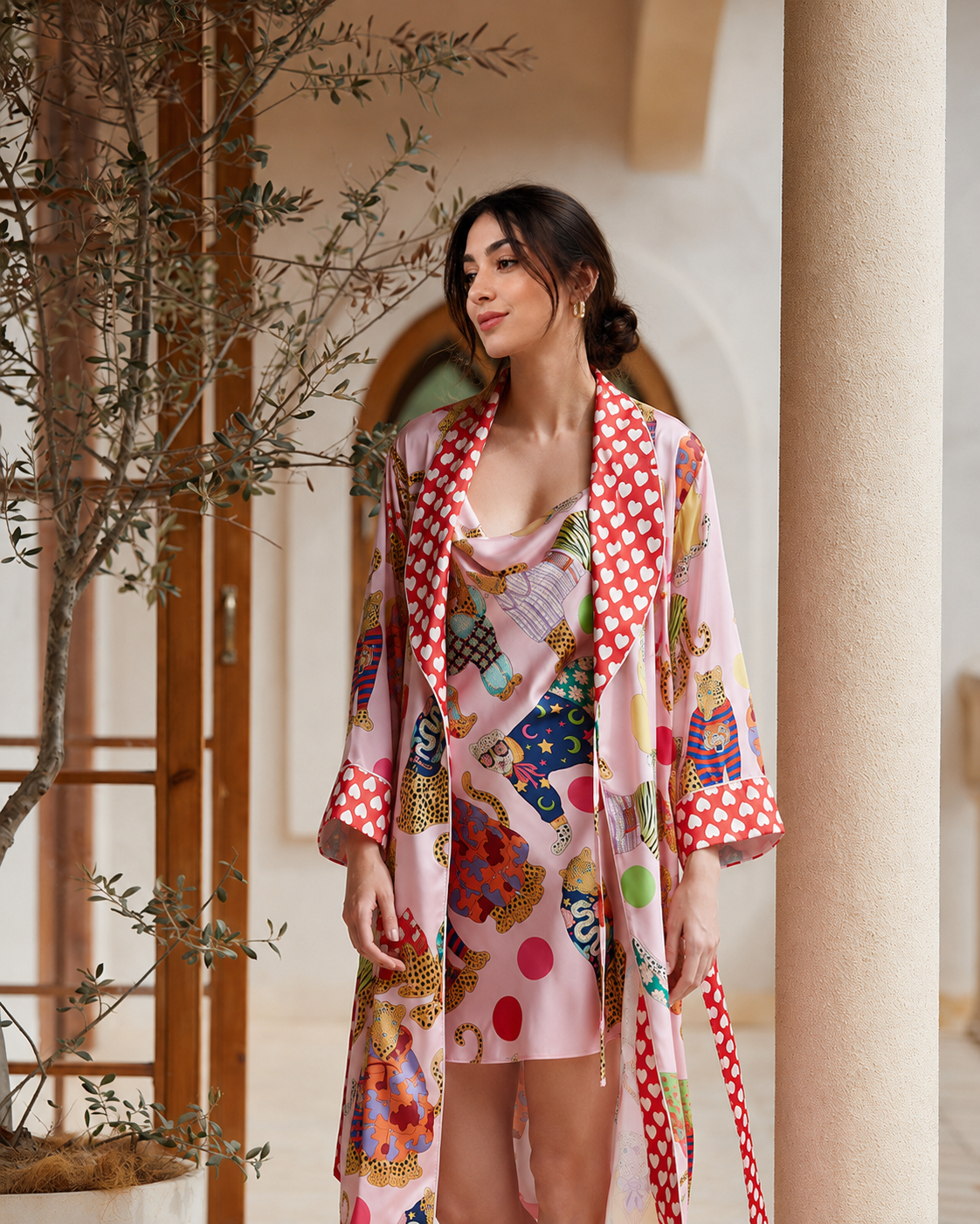 A woman wearing a pink patterned robe and matching dress stands in a warm-toned architectural setting with columns and a tree, featuring a multicolor print with red polka dots and whimsical illustrations.