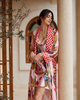 A woman wearing a light pink silk-like robe with a red and white polka-dot collar and a multicolor baroque print, standing in a sunlit courtyard next to a beige column.