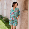 A woman stands outdoors against a beige stucco wall wearing a matching two-piece pajama set with a light aqua background and a colorful flamingo and floral print, plus green and white striped trim.