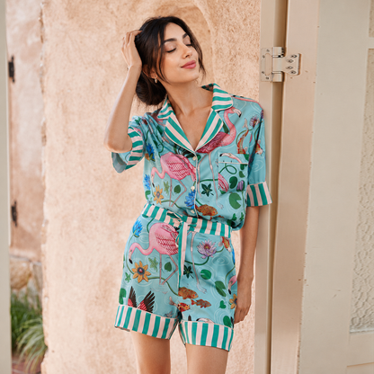 A woman models a two-piece pajama set featuring a tropical flamingo print on a light teal background with green and white striped trim, posed against a rustic beige stucco wall.