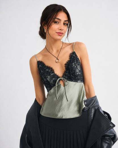 A woman models a two-tone camisole top with a black lace upper section and a sage green satin lower section, styled with a black pleated skirt and a black leather jacket against a light gray background.