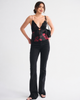 A woman modeling a black camisole top with a red floral pattern and black lace trim, paired with black flared jeans and a small black shoulder bag, against a light gray studio background.