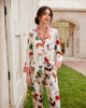 A woman stands outdoors wearing a matching long-sleeve pajama set with a festive Christmas print featuring foxes, Santa Claus, and holiday motifs on an off-white background. The pajamas have red and white striped collars and cuffs.