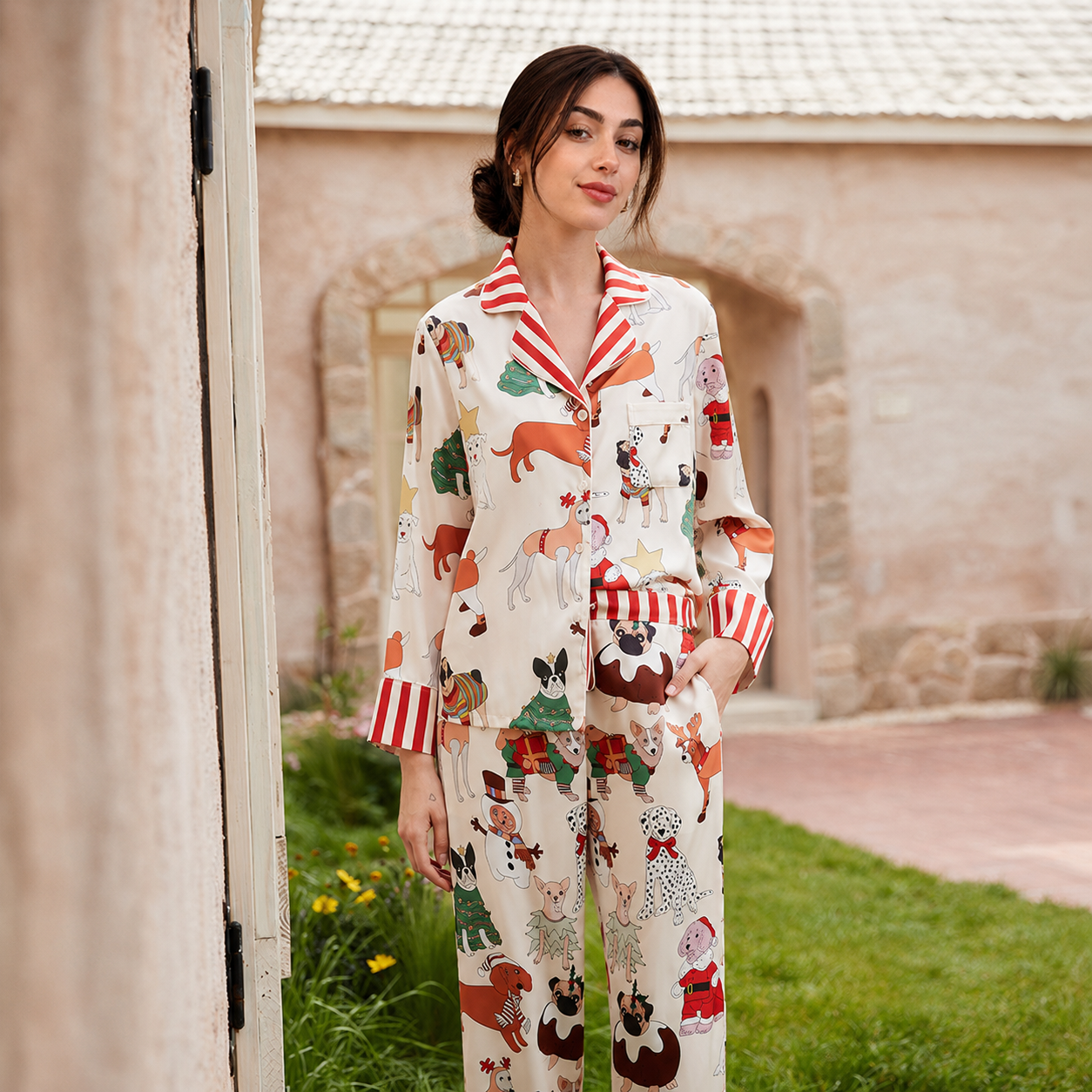 A woman stands outdoors wearing a two-piece pajama set with a cream background and a colorful cartoon dog print, featuring red-and-white striped collar and cuffs. The setting includes a stone building, grass, and a doorway on the left.