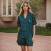 A woman stands outdoors wearing a matching short-sleeve button-front pajama shirt and shorts set in dark teal with thin white vertical pinstripes and a small embroidered animal logo on the chest.