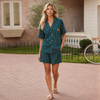 A woman modeling a matching two-piece pajama set with dark teal and white vertical pinstripes, featuring a short-sleeve button-up shirt and shorts, with a small embroidered polo player logo on the chest. She is walking on a brick patio in front of a house.