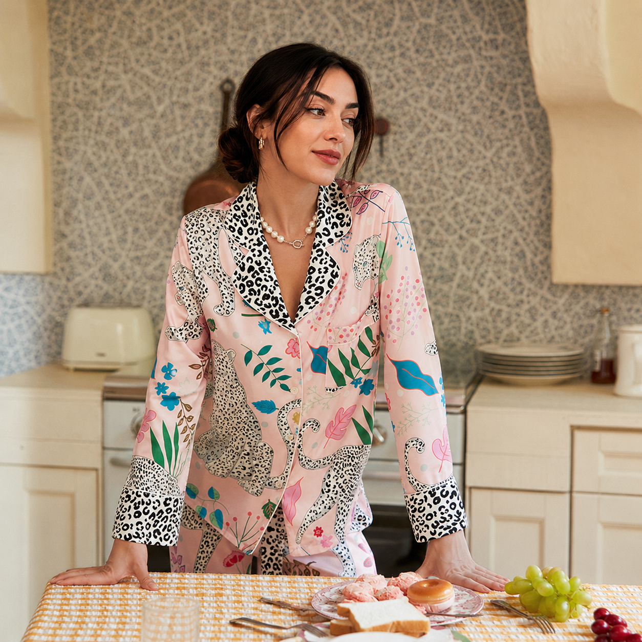 A woman wearing a pink long-sleeve pajama shirt with a multicolor animal and floral print, leaning on a table in a kitchen setting.