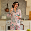 A woman stands in a kitchen wearing a pink patterned pajama set with leopard-print trim. The pajamas feature a colorful botanical and animal print, and she is holding a plate with a piece of cake.