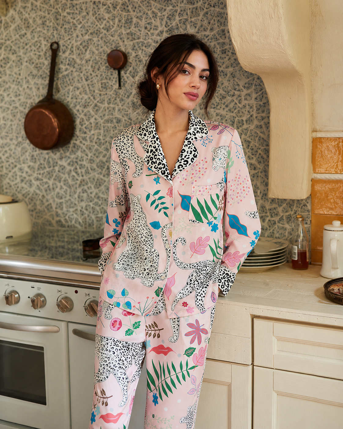 A woman wearing a light pink pajama set with a colorful tropical and leopard print pattern, standing in a kitchen setting.
