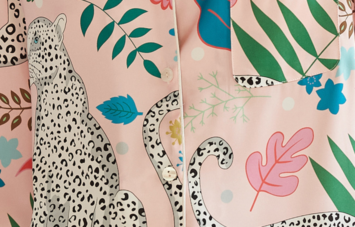 A close-up of a pink button-front shirt or jacket featuring a tropical and safari print with a large spotted leopard, palm leaves, blue flowers, and green foliage.