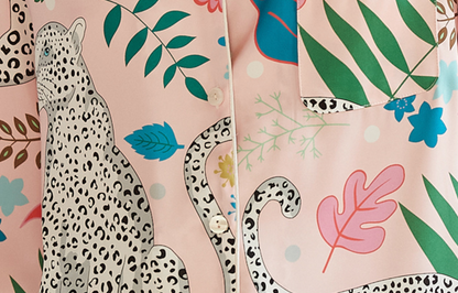 A close-up of a pink button-front shirt or jacket featuring a tropical and safari print with a large spotted leopard, palm leaves, blue flowers, and green foliage.