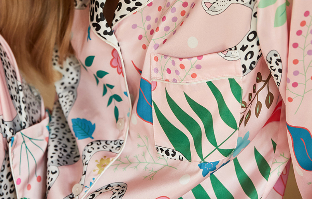 A close-up of a person wearing a light pink satin pajama shirt with a colorful botanical and animal print, featuring green leaves, blue flowers, and black-and-white leopard spots.