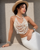 A woman wearing a straw hat, a white multi-strand beaded halter crop top, and a white skirt, posing against a textured gray wall with strong sunlight and shadows.