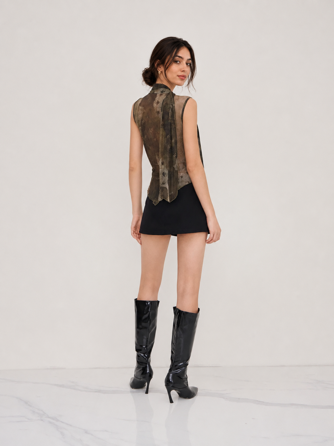 A woman stands with her back to the camera wearing a sleeveless black mini dress with a brown and black patterned high-neck overlay, paired with black knee-high heeled boots, against a plain white studio background.