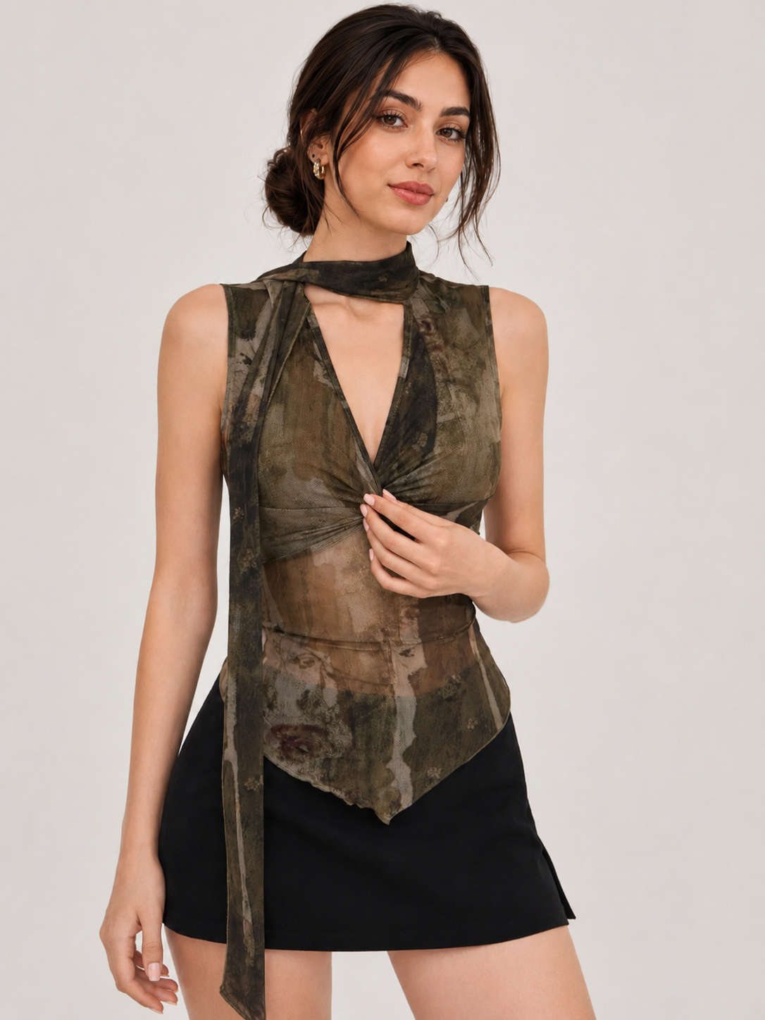 A woman modeling a sleeveless, high-neck wrap top with a distressed camouflage pattern in olive green, brown, and black, paired with a black mini skirt against a light gray studio background.