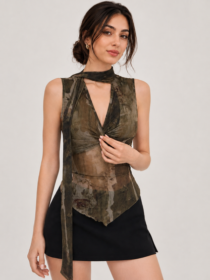 A woman modeling a sleeveless, high-neck wrap top with a distressed camouflage pattern in olive green, brown, and black, paired with a black mini skirt against a light gray studio background.